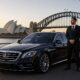 Private driver in Sydney standing beside black luxury sedan with Sydney Opera House and Harbour Bridge in background
