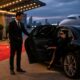 Professional chauffeur assisting a VIP passenger into a luxury black sedan at an exclusive evening event with private jet in the background.