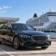 Black chauffeur sedan and suitcases beside a waterfront terminal with a cruise ship in the background.