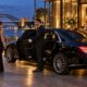 Chauffeur beside a black luxury sedan with formally dressed guests outside a Sydney waterfront awards venue.