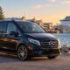 Black Mercedes-Benz V-Class beside a harbour with a cruise ship and heritage buildings at sunset.