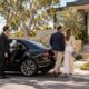 Chauffeur holds a black luxury sedan’s door as a couple approaches a contemporary home for a property inspection.